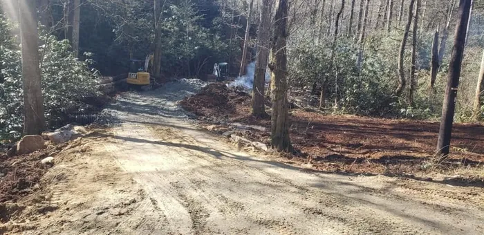 Land clearing in progress with an excavator and a burn pile beside a new gravel drive